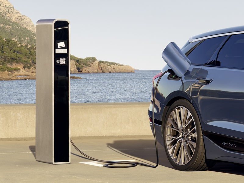 Der S6 Avant e-tron steht rückwärts auf einem Parkplatz und ist dabei an eine Ladestation angeschlossen. Hinter dem Parkplatz sind das Meer und Felsen zu erkennen.