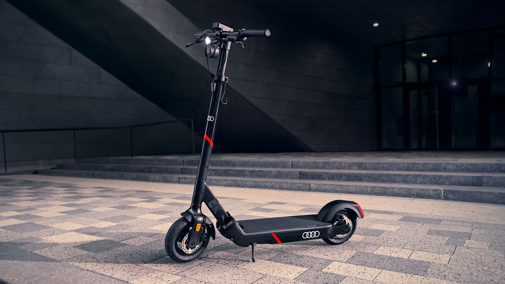 Une trottinette électrique Audi noire avec des accents rouges sur un parvis pavé.