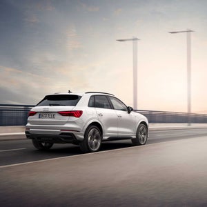 A grey Audi SUV is parked at a charging station beside a modern building. The scene suggests innovation and sustainability against a scenic backdrop.