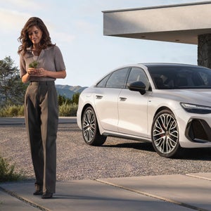 A woman smiles holding a small plant, standing on a sunlit path beside a white Audi. The background shows a modern building and distant mountains.