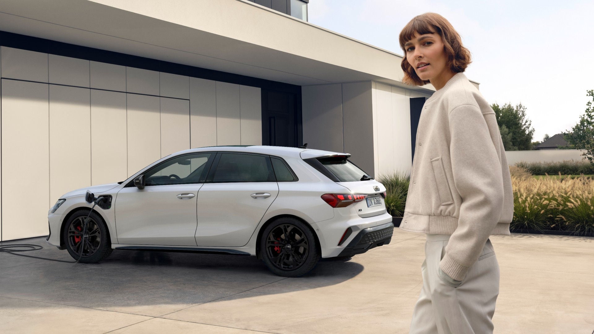 A woman in a light sweater stands near a white electric car charging in a modern driveway.