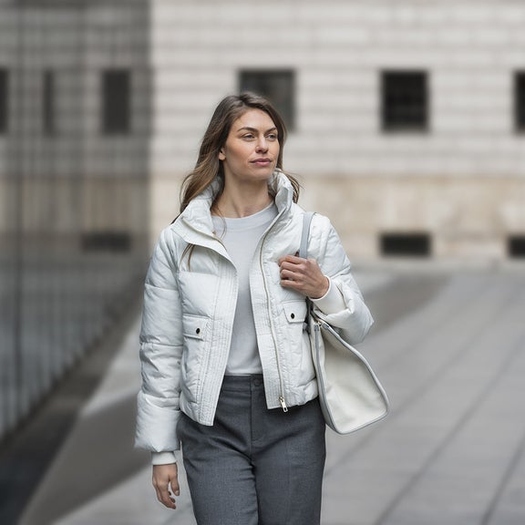 A woman walks past a mirrored building. She is carrying a white leather bag over her shoulder.