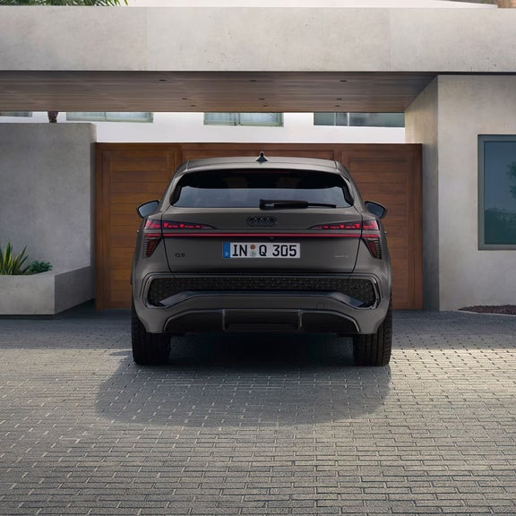 The rear of a grey Audi Q3 Sportback, parked on a paved driveway in front of a stylish house with wooden doors and minimalistic design.