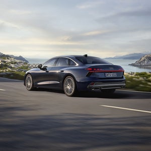 Die A6 Limousine in einer schrägen Heckperspektive, während sie auf einer kurvigen Straße entlang eines Fjords fährt. 