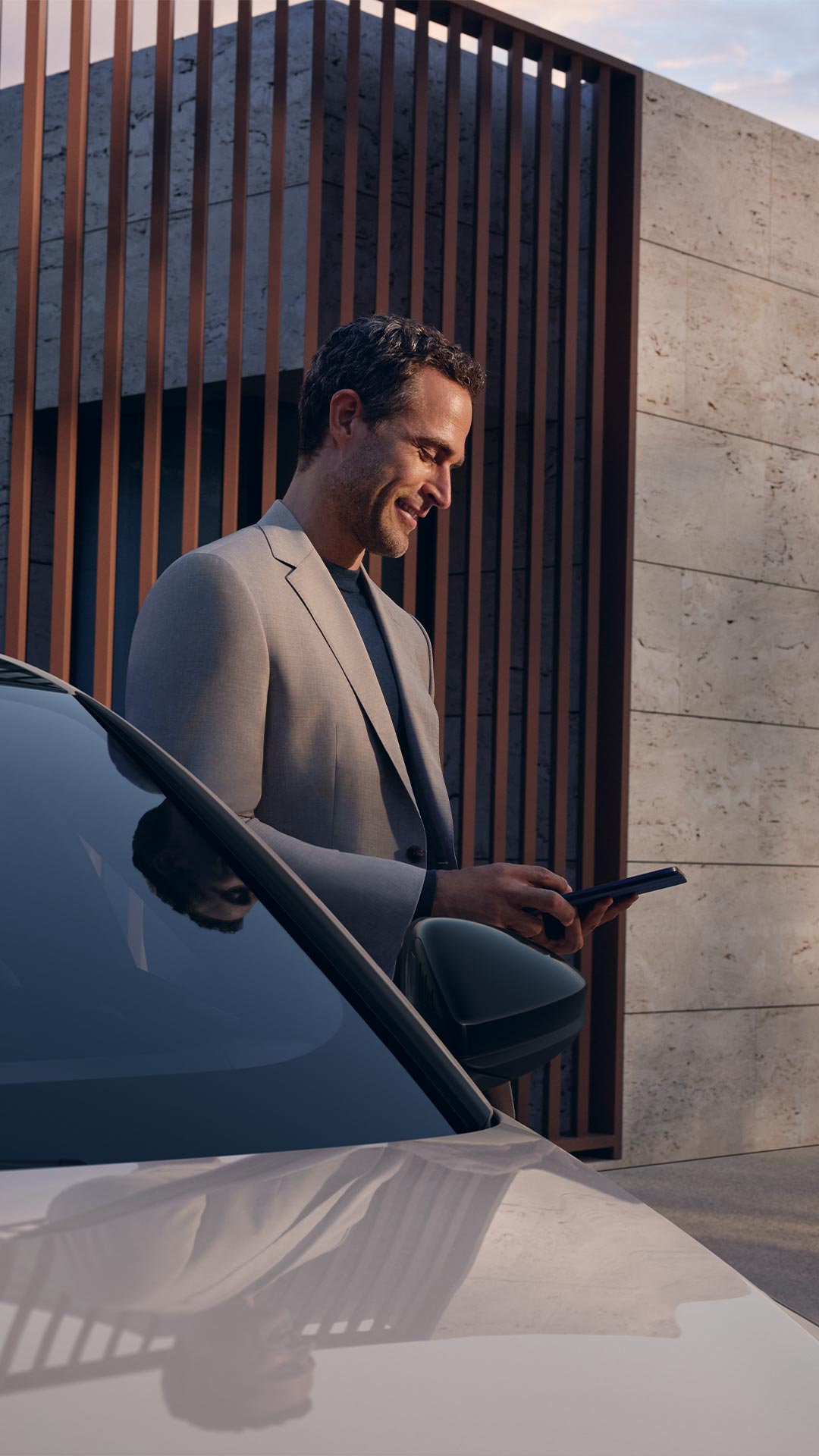 Mann in grauem Anzug steht neben einem Audi und blickt lächelnd auf ein Smartphone, im Hintergrund moderne Hausfassade.