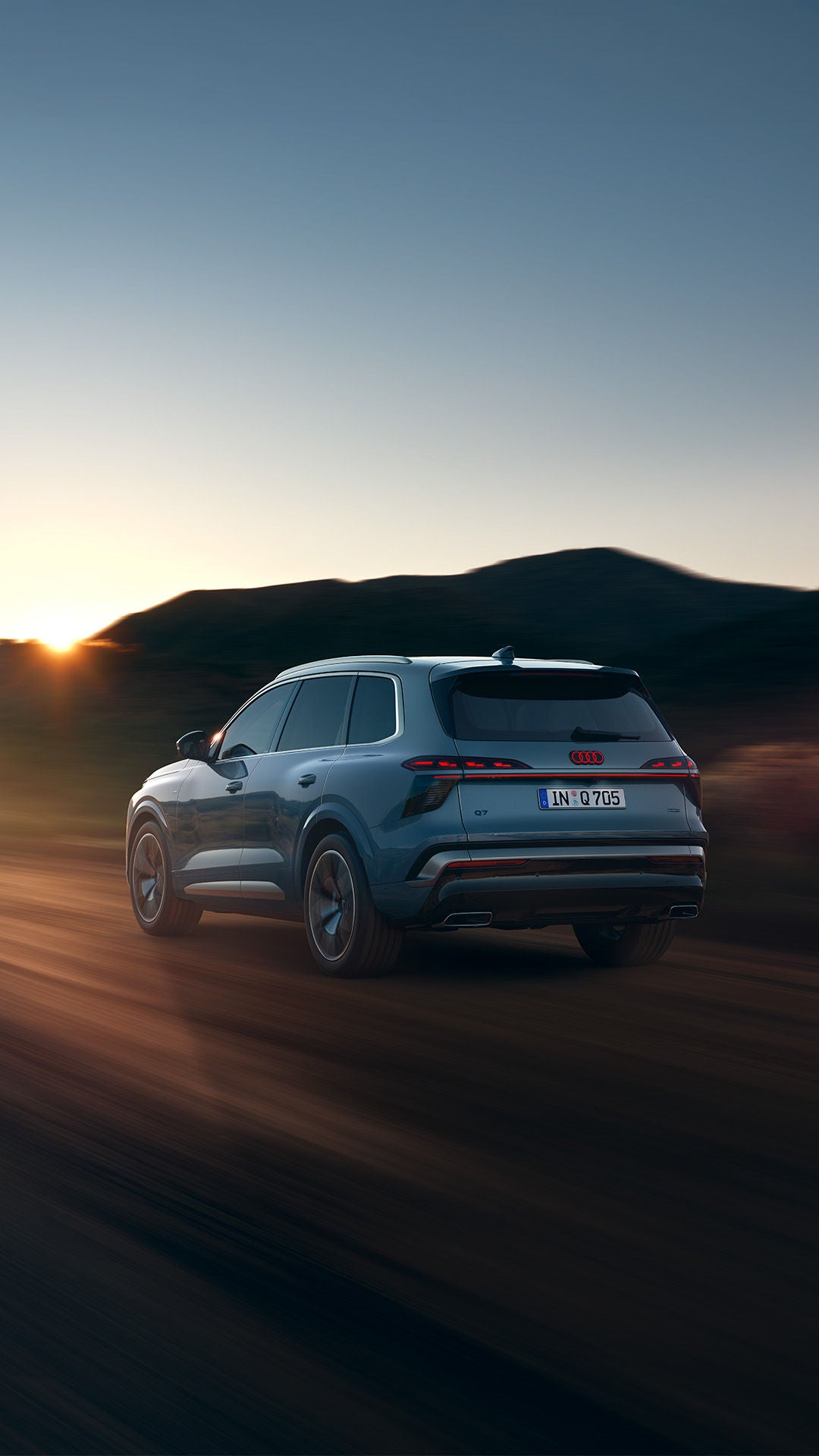 Der neue Audi Q7 in Dreiviertel-Heckansicht fährt auf einer Landstraße in den Sonnenuntergang.