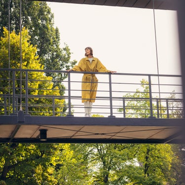 Frau in einem gelben Mantel steht auf einer Brücke und hält sich am Geländer fest und schaut in die Natur. Im Hintergrund sind Bäume mit grünen Blättern