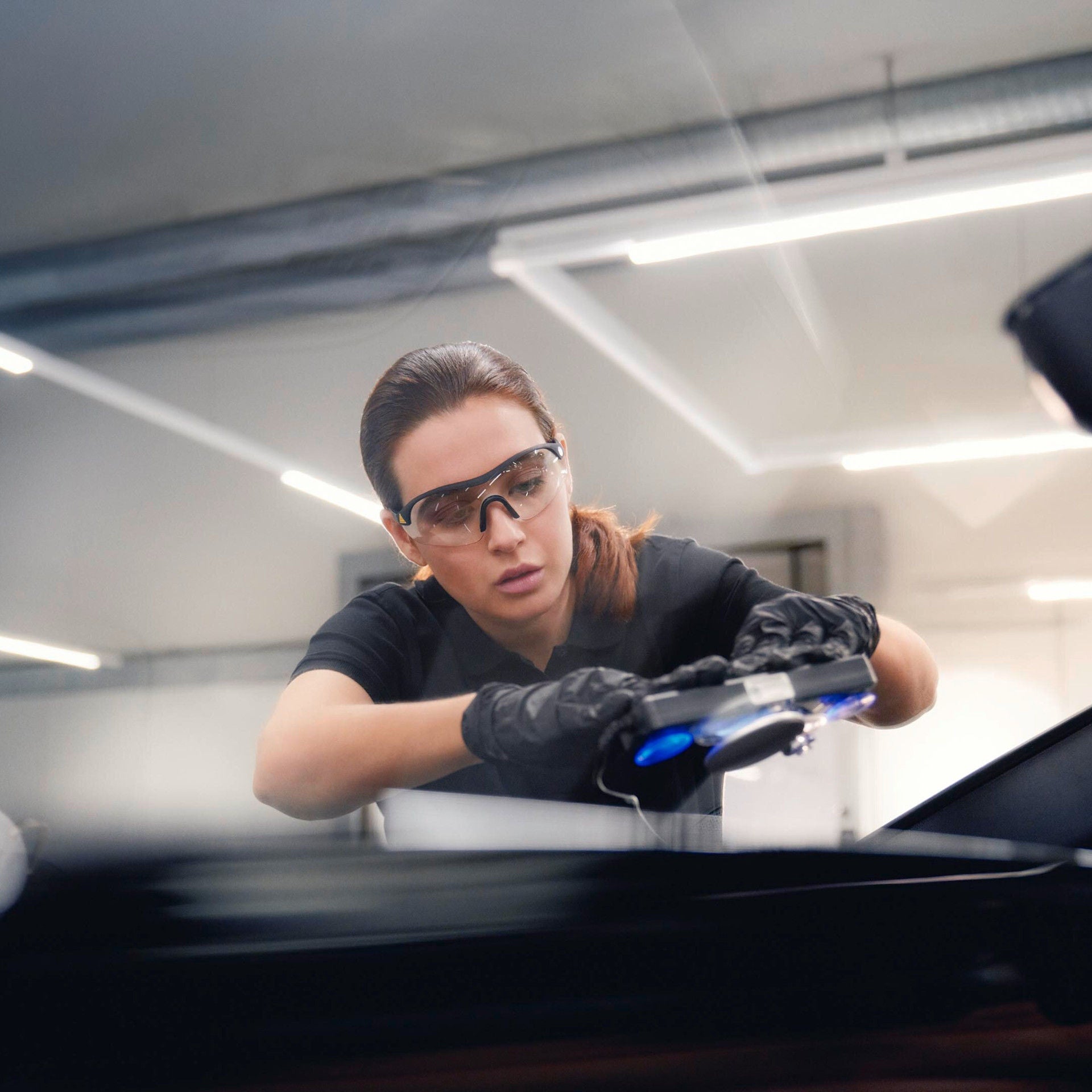 Mitarbeiterin mit Schutzbrille und Handschuhen arbeitet mit einem Prüfgerät an einem Auto.