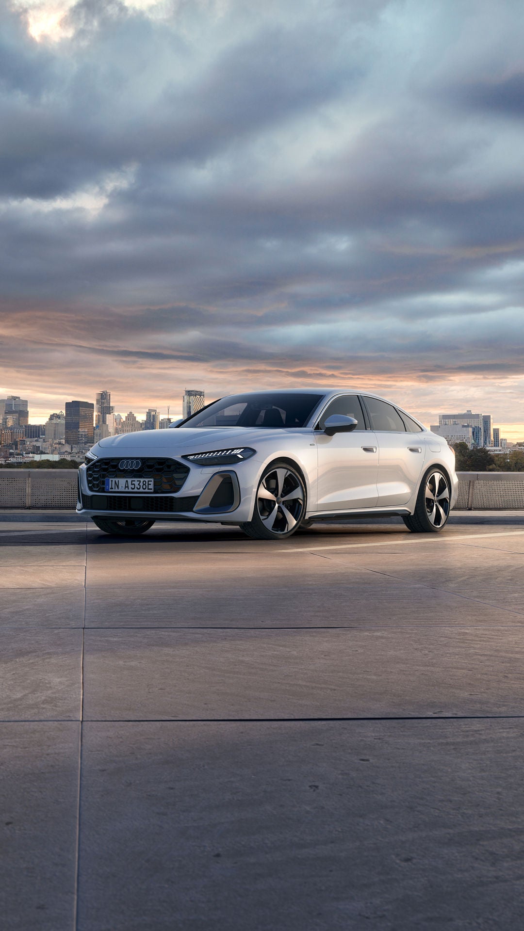 A sleek white Audi A5 Saloon e-hybrid car is parked on a rooftop overlooking a city skyline at sunset.