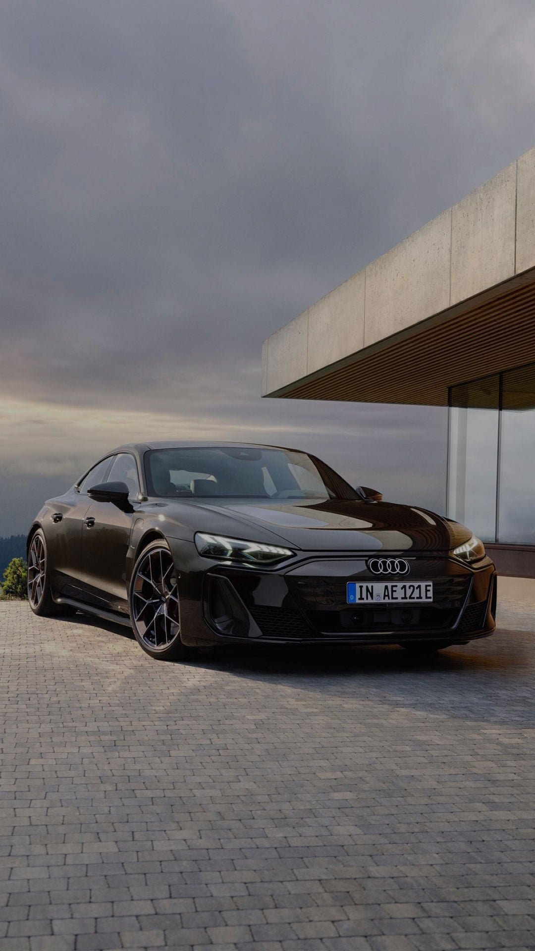 Black Audi parked on a modern stone driveway, beside a sleek glass-walled building. Scenic hills under a cloudy sky create a serene backdrop.