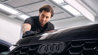 Mann mit Handschuhen poliert die schwarze Motorhaube eines Audi in heller Werkstatt.