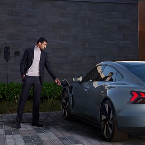 A man in a suit charges a sleek, grey Audi car parked on a modern driveway. The scene conveys a sense of sophistication and eco-friendly living.