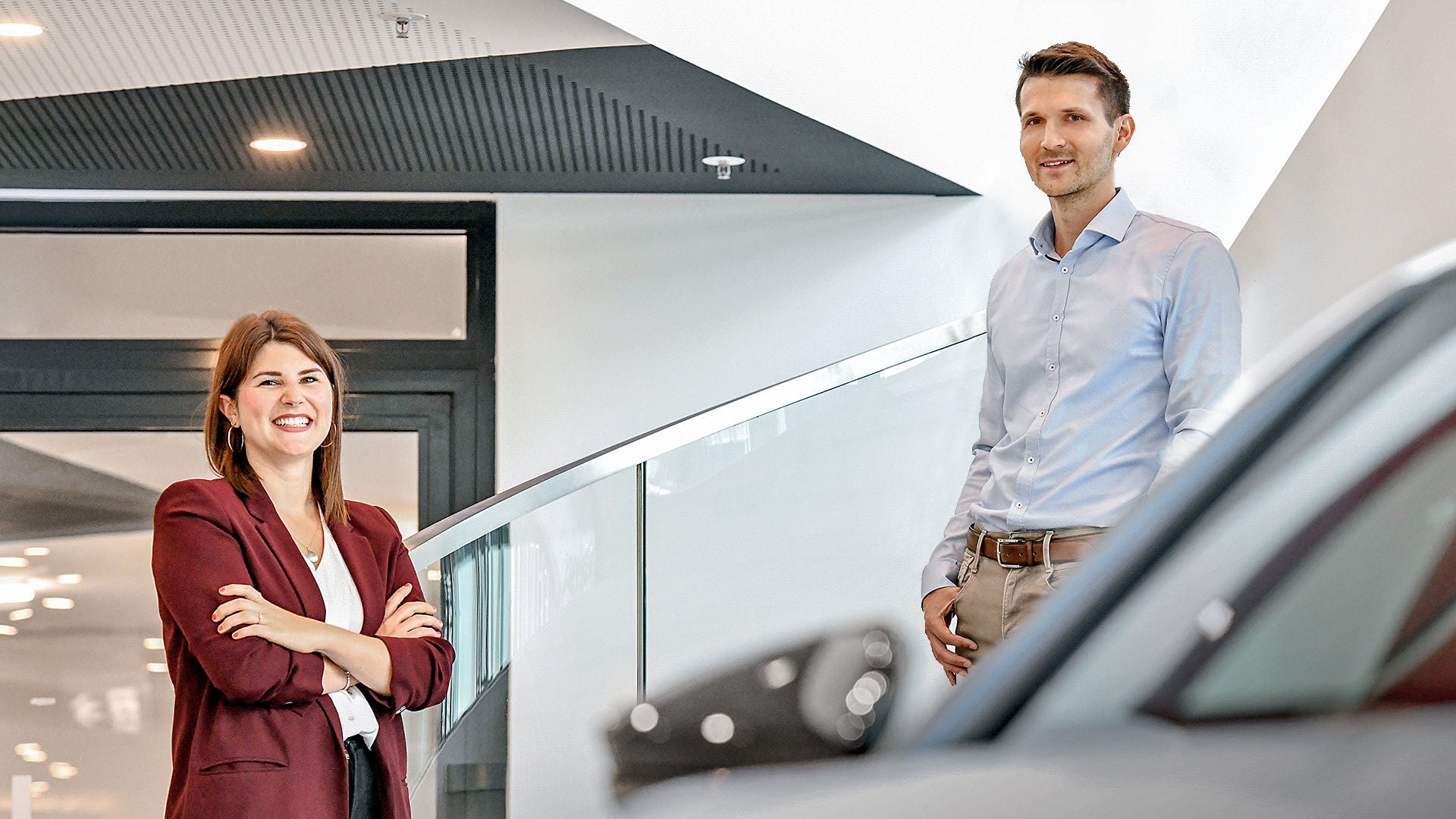 A smiling woman and a man are standing next to a car in a car dealership.