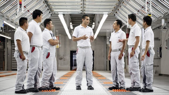 Sechs Audi-Mitarbeiter im weißen Arbeitsoutfit im Gespräch in heller Produktionshalle.