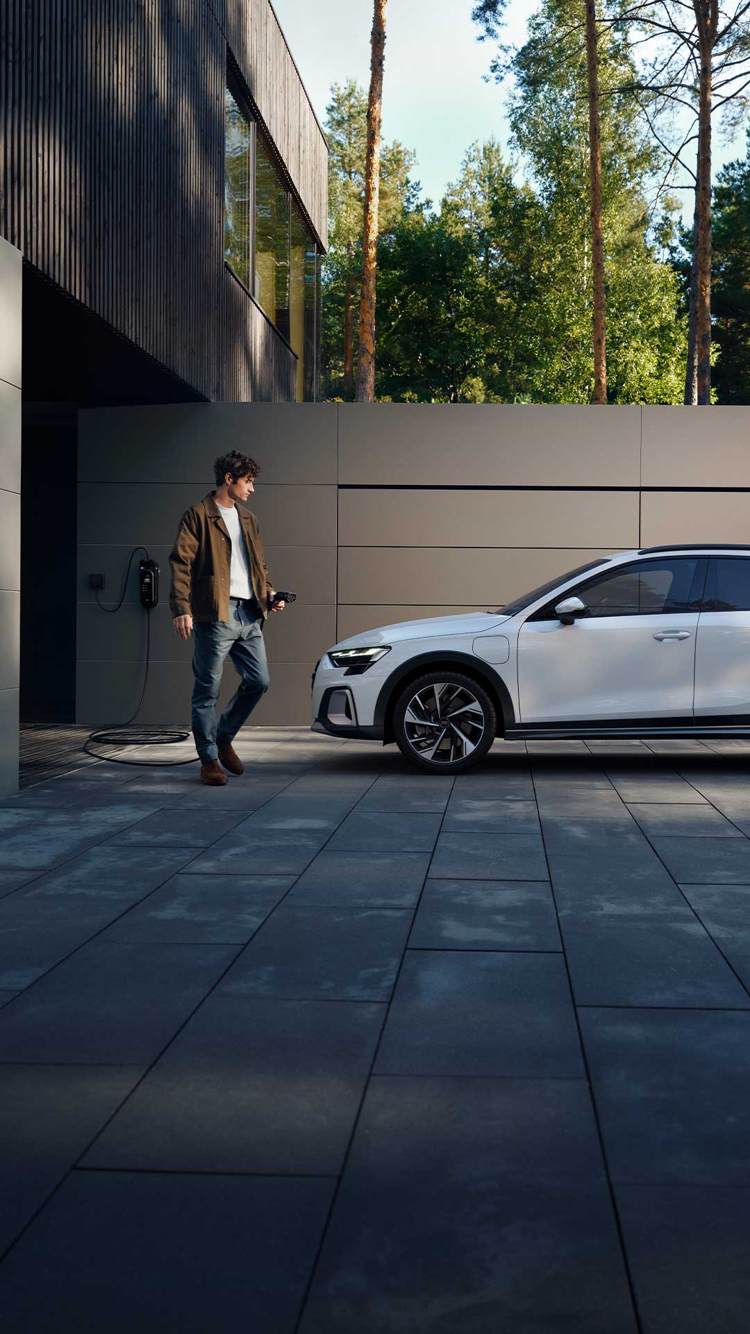Ein Mann mit Ladekabel steht vor einem weißen Audi A3 allstreet TFSI e, der vor einer modernen Wand geparkt ist.