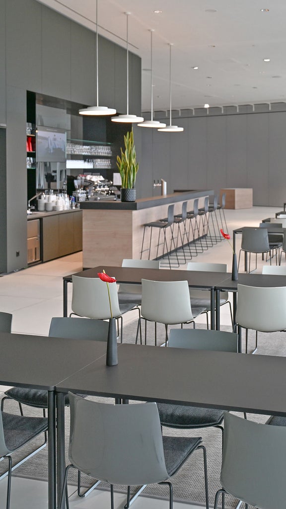 Heller Cafébereich mit Kaffeetheke, Getränkekühlschränken und zahlreichen Tischen und Stühlen. Bodentiefe Fenster lassen Tageslicht in den Raum.