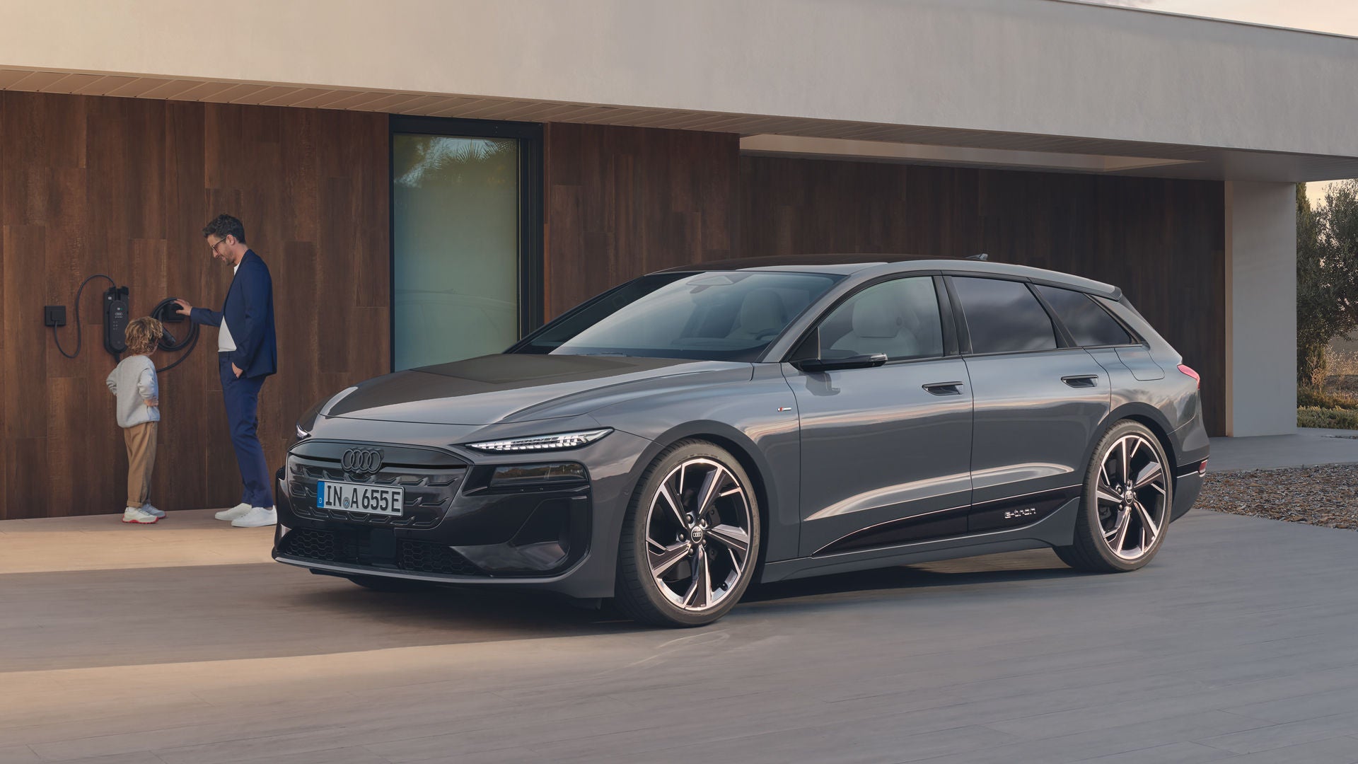 De Audi A6 Avant e-tron staat geparkeerd voor een modern huis. Aan de muur van het huis is een laadstation gemonteerd, waar een vader met zijn kind staat.