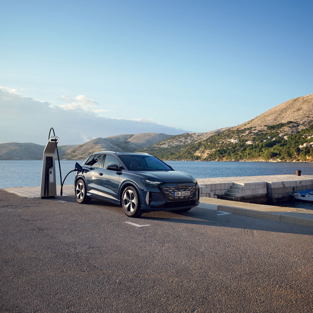 Audi Q4 e-tron bleue en recharge sur une borne électrique au bord de l’eau, avec un quai, un bateau et des collines en arrière-plan sous un ciel dégagé.