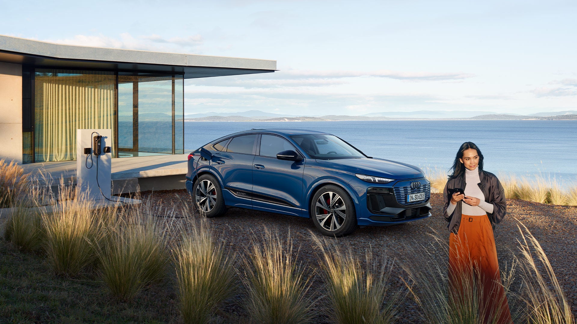 Frau steht vor einem blauen Audi SQ6 Sportback an einer Ladestation am modernen Haus mit Meerblick, umgeben von Graslandschaft.