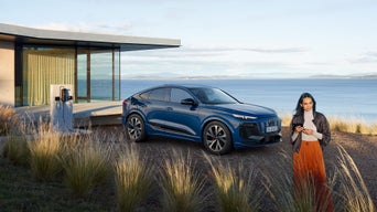 Eine Frau steht neben einem blauen Audi e-tron im Hintergrund ein modernes Haus am Wasser.