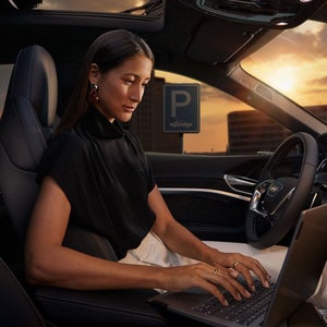 Frau arbeitet bei Sonnenuntergang mit Laptop auf dem Fahrersitz eines geparkten Audi, im Hintergrund ein Parkschild.