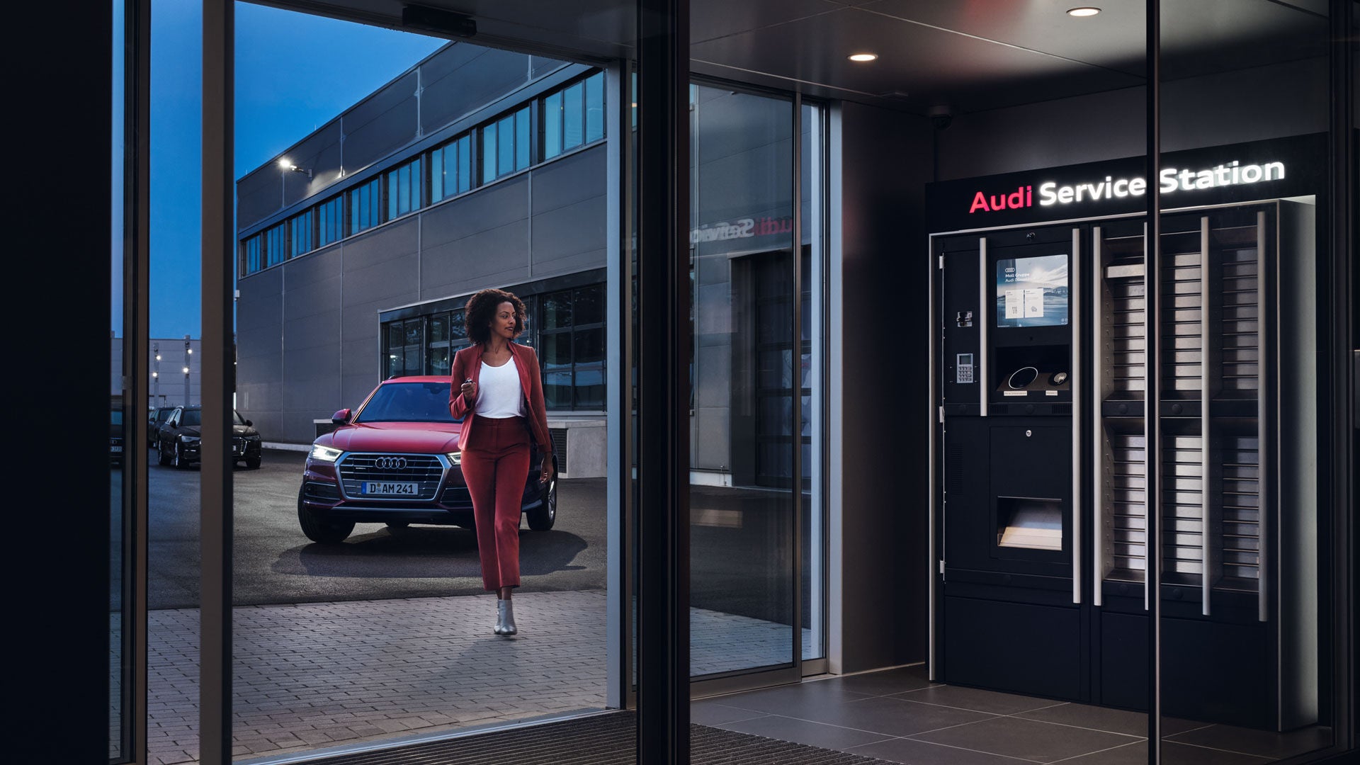Frau geht am Abend zu einer Audi Service Station, ein roter Audi steht vor dem Gebäude.