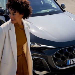 Frau in beiger Jacke steht neben einem Audi Q5, aufgenommen von schräg oben, mit Fokus auf Fahrzeugfront und Kühlergrill.