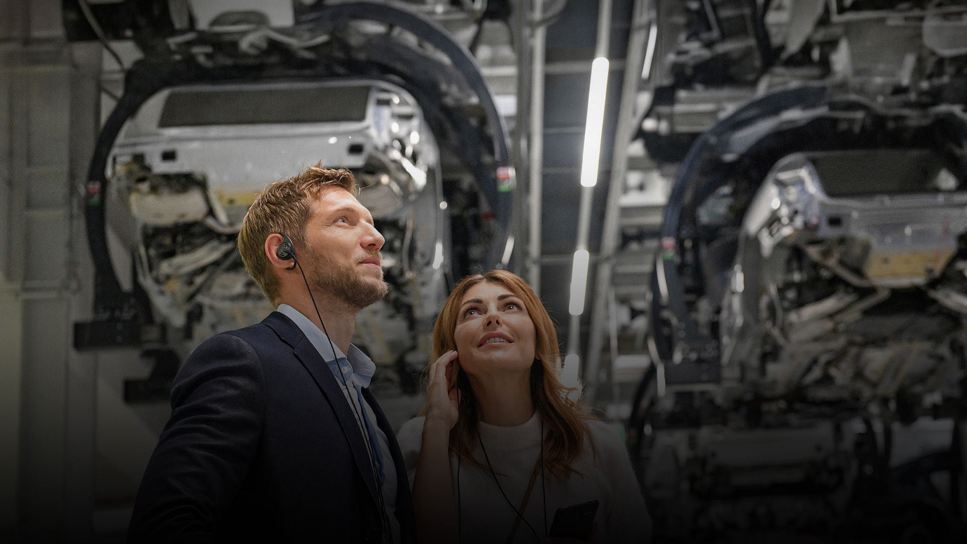 Ein Mann und eine Frau nehmen an einer Erlebnisführung teil und betrachten die Karosserie eines Audi Modell von unten.