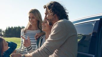 A family enjoys a sunny day outdoors near a car, sharing a joyful moment and engaging in conversation. Green trees are in the background.