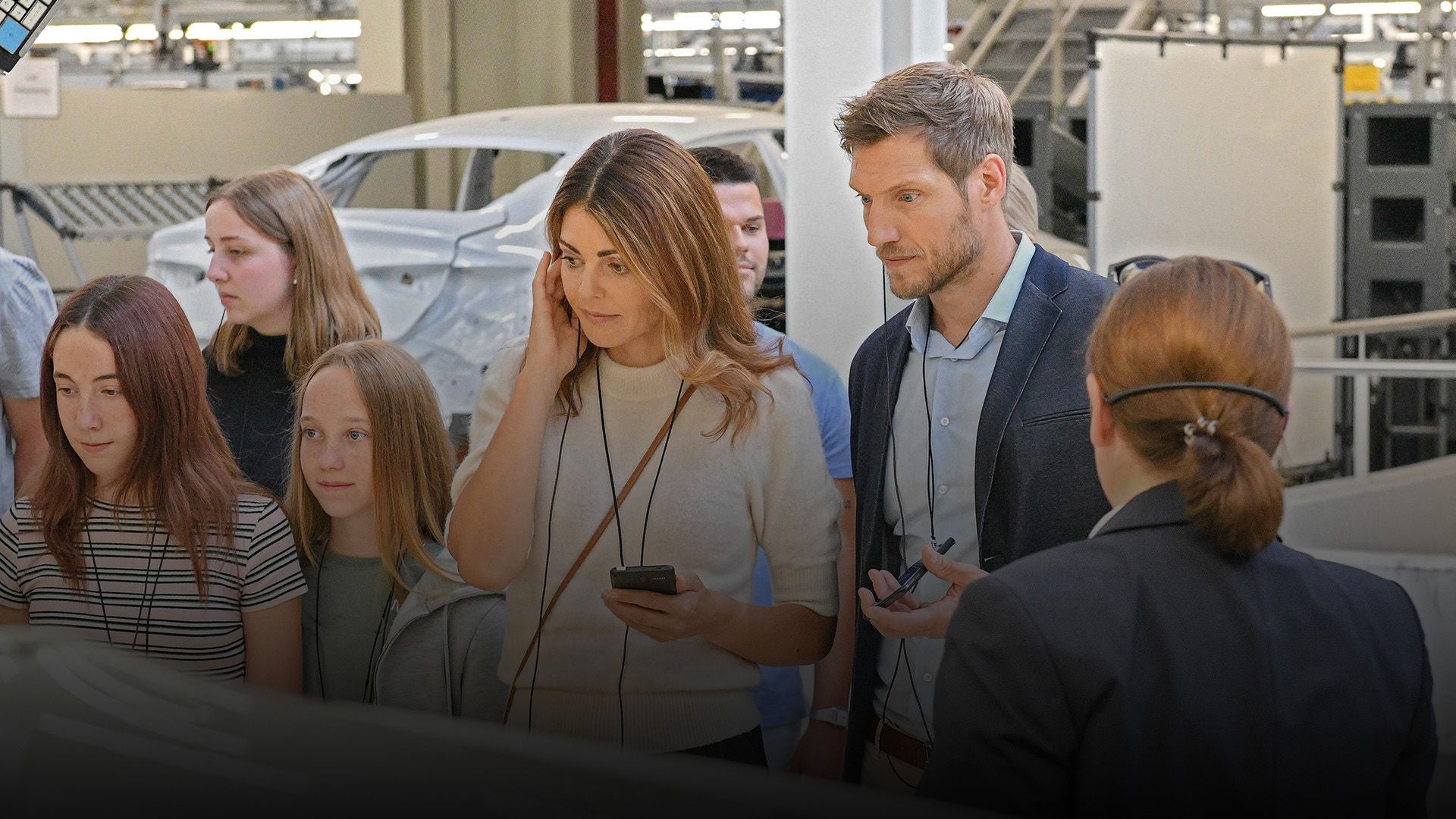 Group of people on a factory tour, man and woman in the foreground with headsets in the production hall.