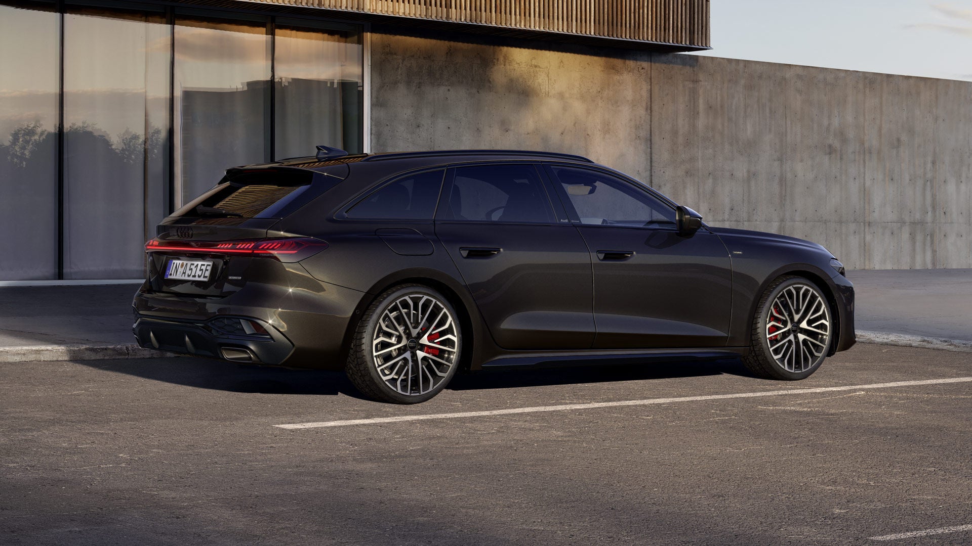 A5 Avant e-hybrid i lackeringen Havana Black Metallic står i en vinkel på en parkeringsplats framför en byggnad.