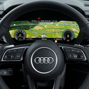 Black Audi steering wheel with silver logo, behind which is a digital display showing a map, speedometer and other car information.