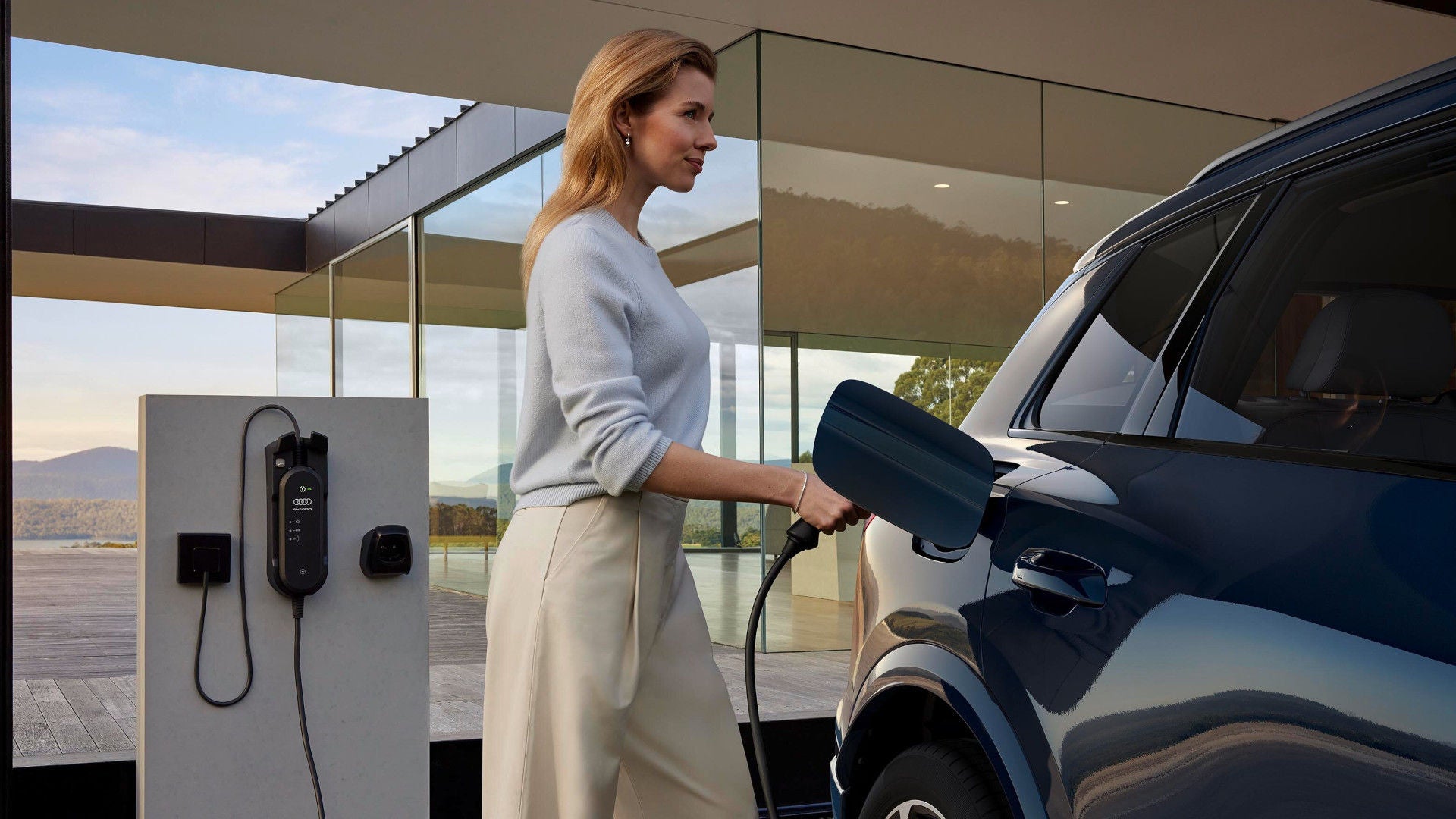 Frau lädt ein Elektroauto an einer modernen Ladesäule vor einem verglasten Gebäude.