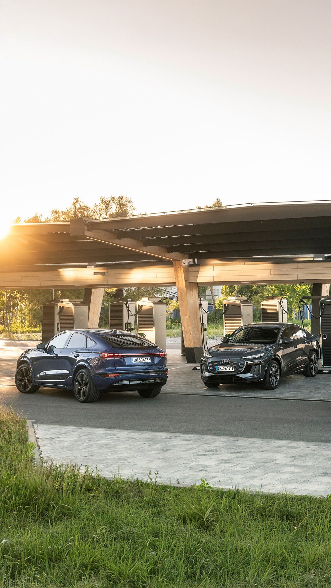 Three Audi Q6 Sportback e-tron cars at an Audi charging hub. One is charging while the others drive to the charging stations under the beige roof structure.