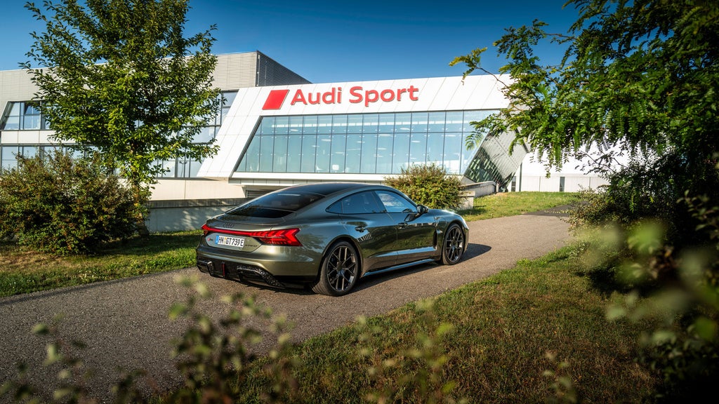 Ein grüner Audi RS e-tron GT auf einer Auffahrt, im Hintergrund die Firmenzentrale mit Logoschriftzug der Audi Sport GmbH.
