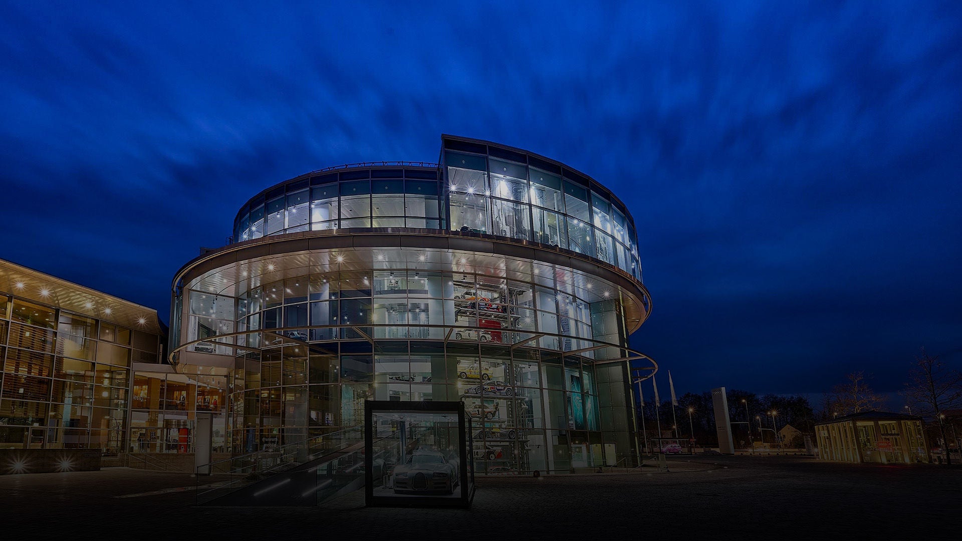 Round, modern building with glass façade shines at night under a cloudy, blue sky.