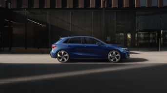 Une Audi A3 Sportback bleue garée devant un immeuble moderne à la façade vitrée, dans un environnement urbain. 