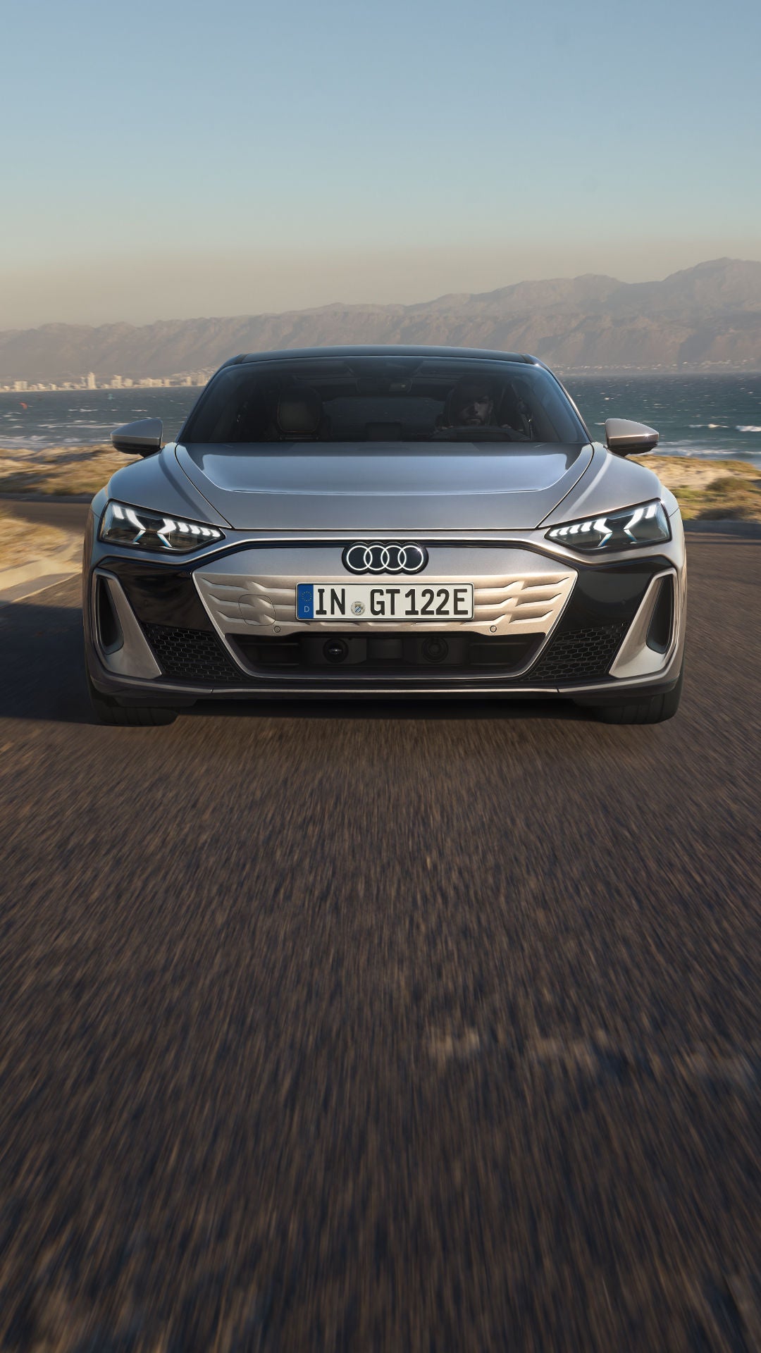 Ein silberner Audi S e-tron GT steht auf einer gepflasterten Auffahrt und ist an eine Ladestation angeschlossen. Im Hintergrund sind moderne Häuser und eine Dünenlandschaft zu sehen.