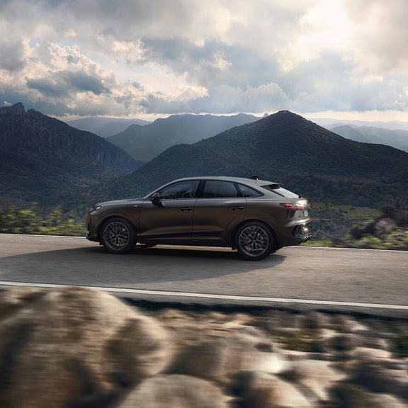 A matte black Audi Q3 driving along a mountain road with scenic green hills and cloudy sky in the background.