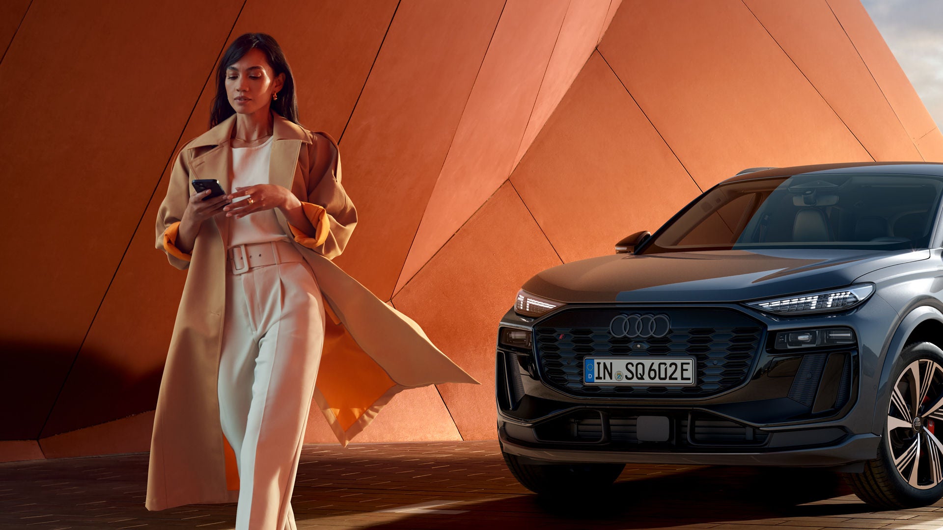 Frau steht lächelnd vor zwei geparkten Audi-Fahrzeugen und bedient ein Smartphone, moderner Holzbau im Hintergrund. 