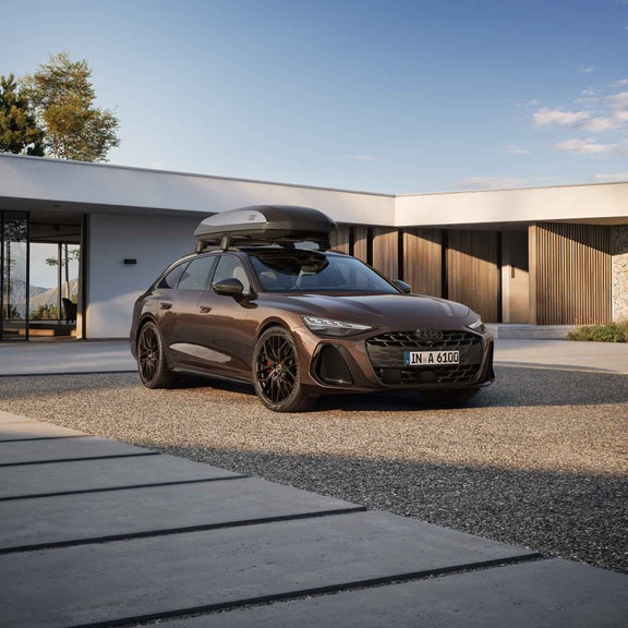 Brauner Audi A6 Avant mit Dachbox steht vor moderner Villa, Mann neben dem Auto und lächelt, Frau und Kind sitzen auf Bank im Hintergrund.