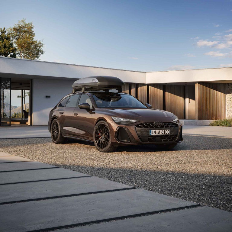 Brauner Audi A6 Avant mit Dachbox steht vor moderner Villa, Mann neben dem Auto und lächelt, Frau und Kind sitzen auf Bank im Hintergrund.