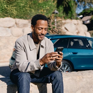 Ein Mann sitzt im Freien vor einer Felslandschaft und blickt auf ein Smartphone, das er mit beiden Händen hält. Hinter ihm steht ein türkisfarbenes Auto.