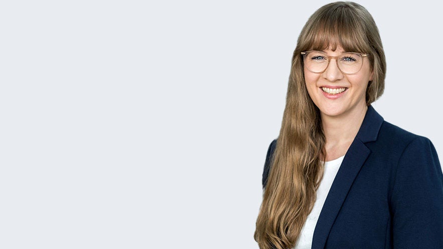 Anne Bodenloher: Porträt lächelnde junge Frau mit langem hellbraunem Haar und großer Brille im Businessoutfit vor hellgrauem Hintergrund.