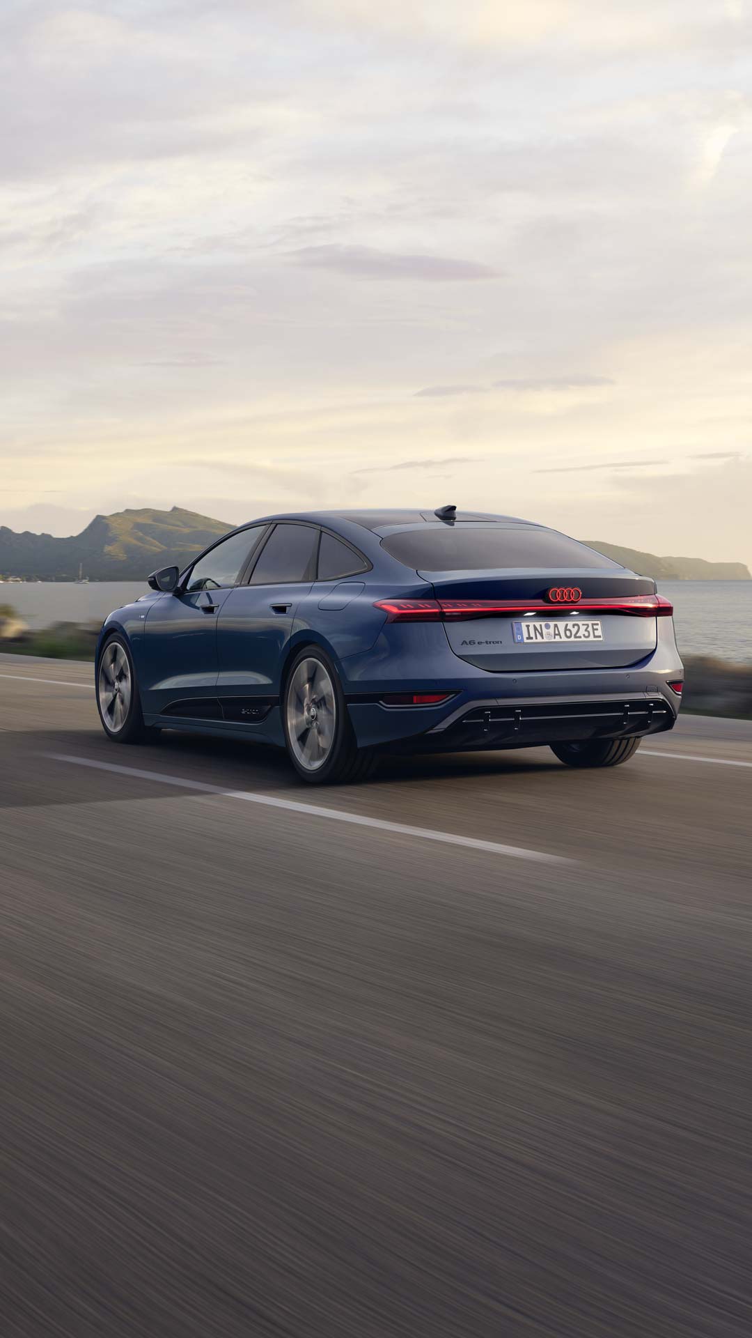 Der A6 Sportback e-tron aus schräger Heckansicht fährt eine Straße parallel zum Ufer entlang. Im Hintergrund sind Berge zu erkennen.&nbsp;