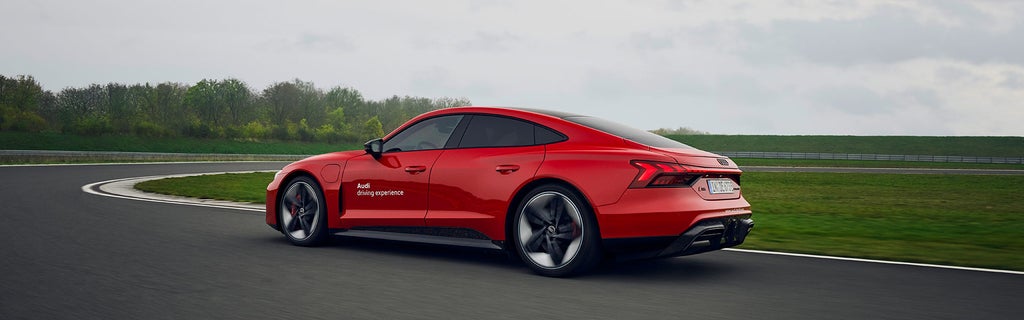 Red Audi RS e-tron GT performance on a racetrack in a curve, rear side view; door with white lettering “Audi driving experience.”