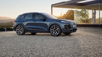Dunkelblauer Audi Q6 SUV e-tron steht auf einem Kiesplatz vor einem modernen Haus, Frau mit Smartphone im Hintergrund.