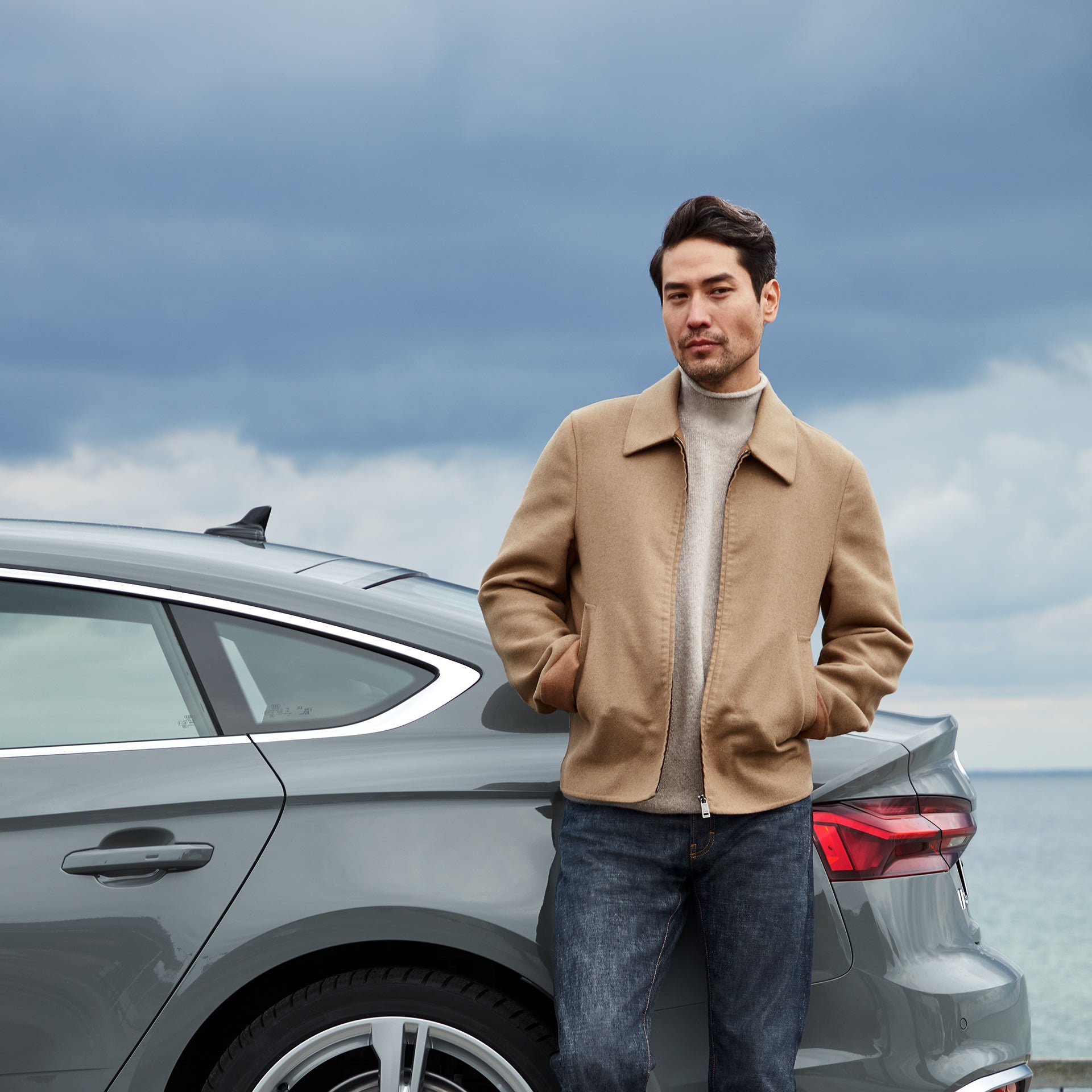 Man standing next to his dark grey Audi which is parked by the sea