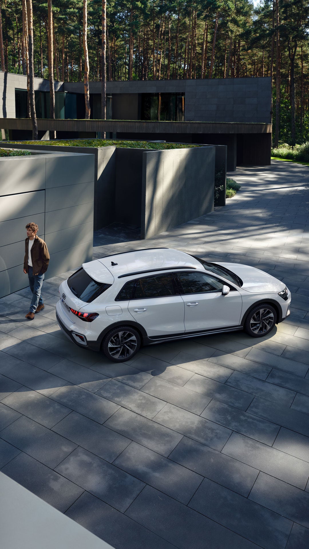 Ein Mann geht neben einem weißen Audi A3 auf einer gepflasterten Auffahrt mit moderner Gebäudefassade entlang.