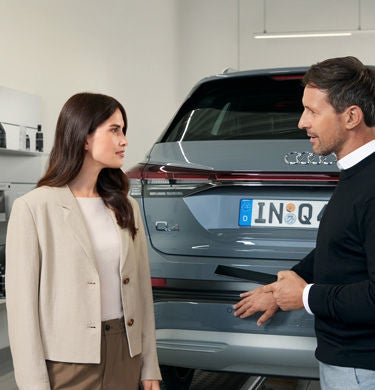 Kundin und Berater sprechen vor dem grauen Audi-Heck im Autohaus; Regale mit Pflegeprodukten im Hintergrund.